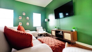 Een kamer met groene wanden, een flatscreen televisie, een witte bank met rode kussens, een klein houten bijzettafeltje met een plant en een raam met jaloezieën.