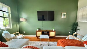 Een moderne woonkamer met een groene wand, een flatscreen televisie boven een houten kast, en een groot raam met jaloezieën.