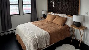Ein Schlafzimmer mit einem Bett, zwei Nachttischlampen, einem großen Fenster mit Vorhängen, einem Deckenventilator und einem weißen, flauschigen Teppich.