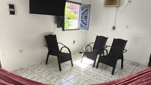 A room with two black wicker chairs, a hammock, a TV mounted on the wall, and a window with a blue shutter. - Renata Fogaça  (Maragogi)