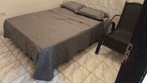 A small, neatly made bed with a checkered bedspread and pillows, a dark wicker chair, and a tiled floor. - Renata Fogaça  (Maragogi)