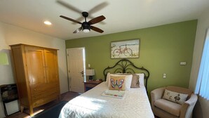 A bedroom with a bed, a wooden wardrobe, a chair, a ceiling fan, and a painting on the wall. - BESO DEL SOL (Huntington Beach)
