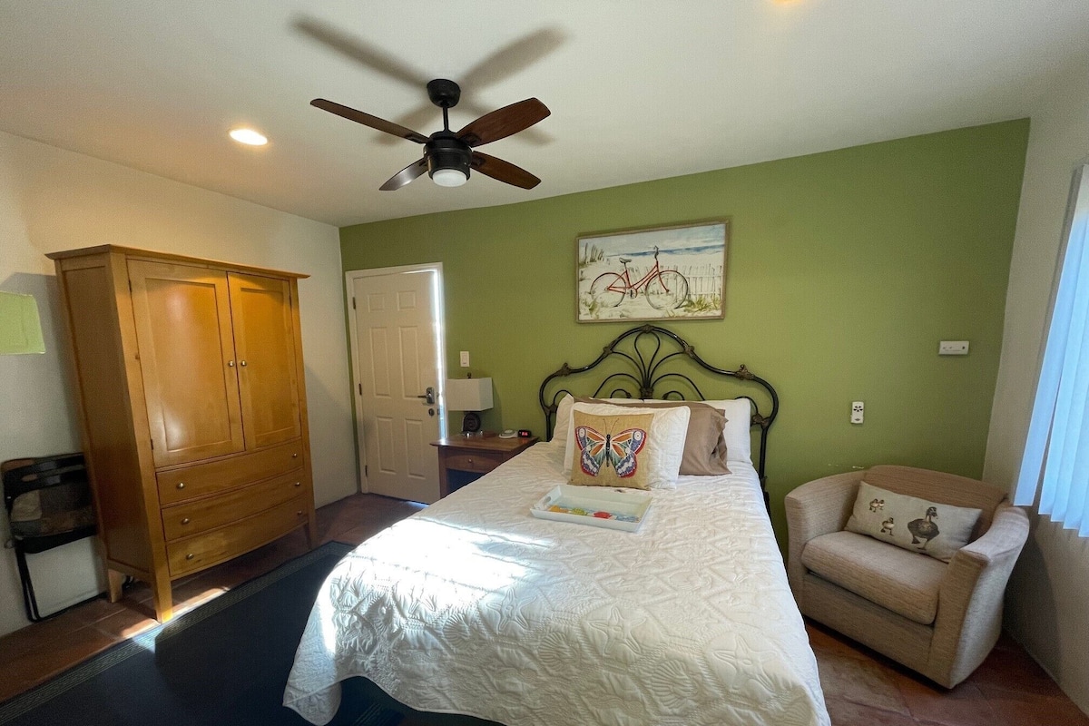 A bedroom with a bed, a wooden wardrobe, a chair, a ceiling fan, and a painting on the wall.