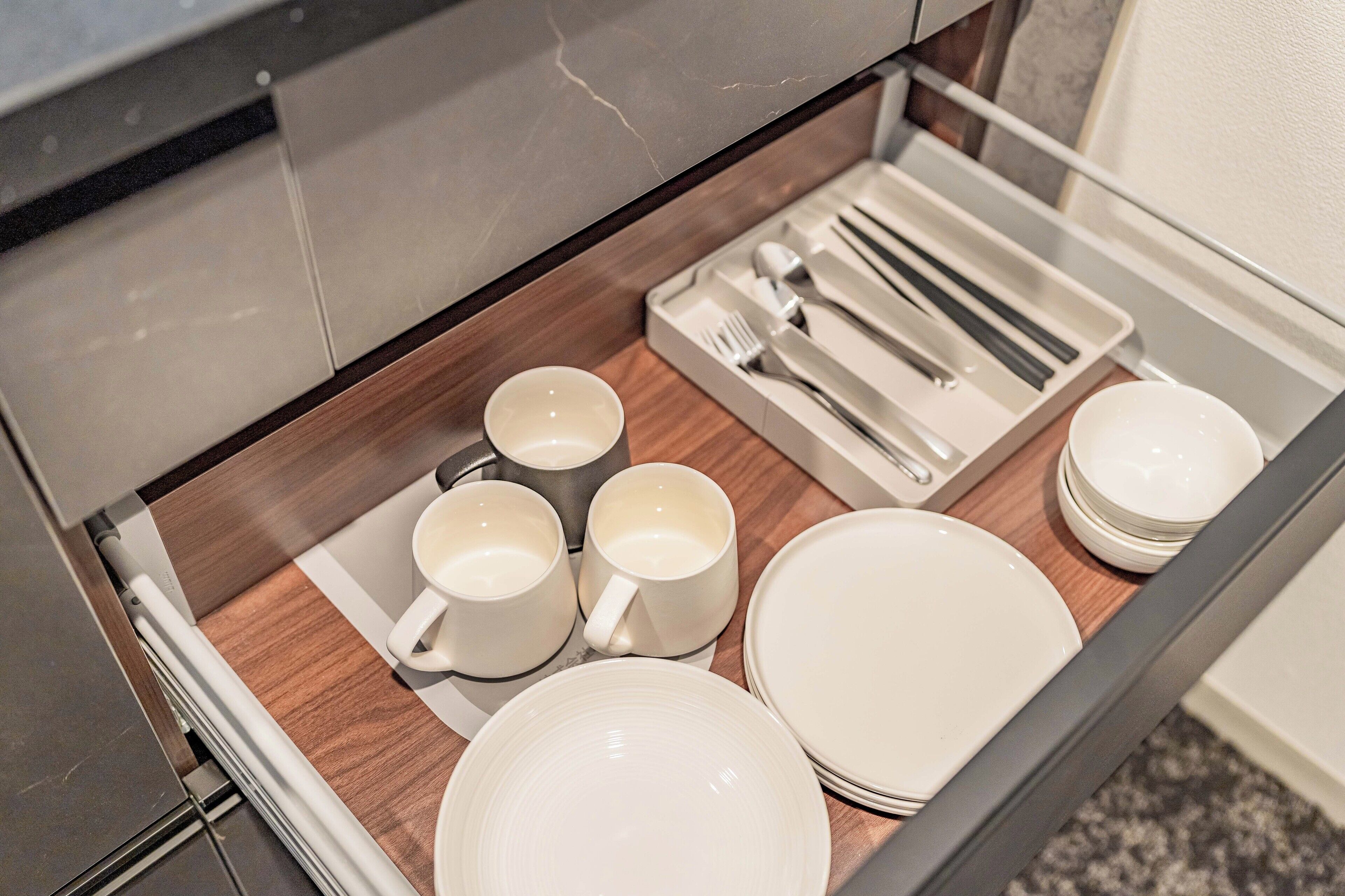A drawer with neatly arranged white mugs, plates, and cutlery.