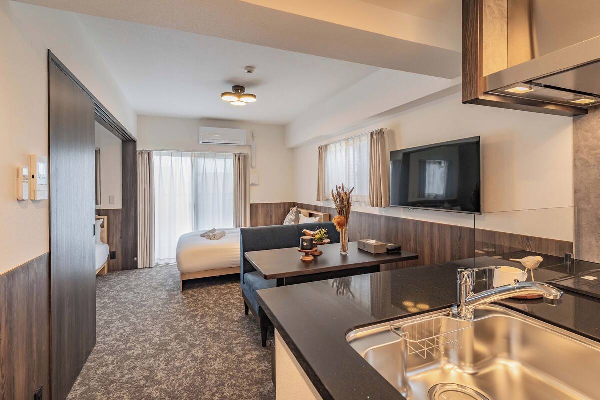 A modern hotel room with a bed, a sofa, a dining table, a kitchen area with a sink, and a flat-screen TV mounted on the wall.