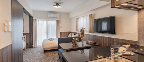 A modern hotel room with a bed, a sofa, a dining table, a kitchen area with a sink, and a flat-screen TV mounted on the wall.