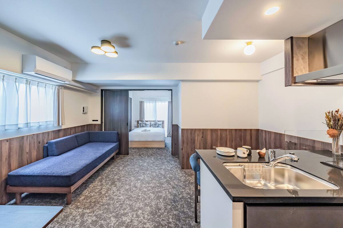 A modern hotel room with a sofa, a bed, a kitchenette, and a bathroom visible through an open door.