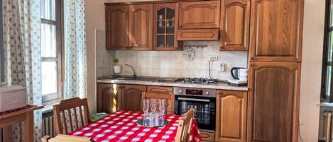 Une cuisine avec des meubles en bois, une cuisinière à gaz, un four et une table à manger recouverte d’une nappe à carreaux rouges et blancs.