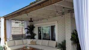 A covered outdoor seating area with a sofa and wicker tables, potted plants, and a wooden deck. - Quiet Laguna Beach Home- 1  Block to Ocean - Pool & Spa-Pets - Games - Sleeps 14 (Panama City Beach)