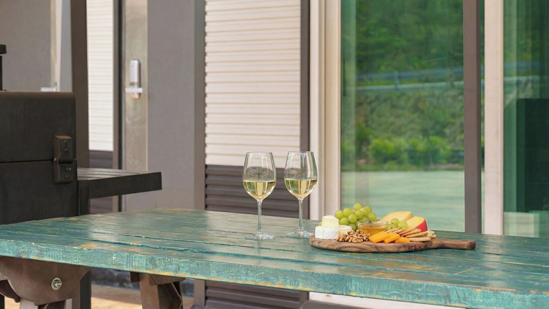 A table with two glasses of white wine, a wooden board with cheese, grapes, nuts, and crackers.