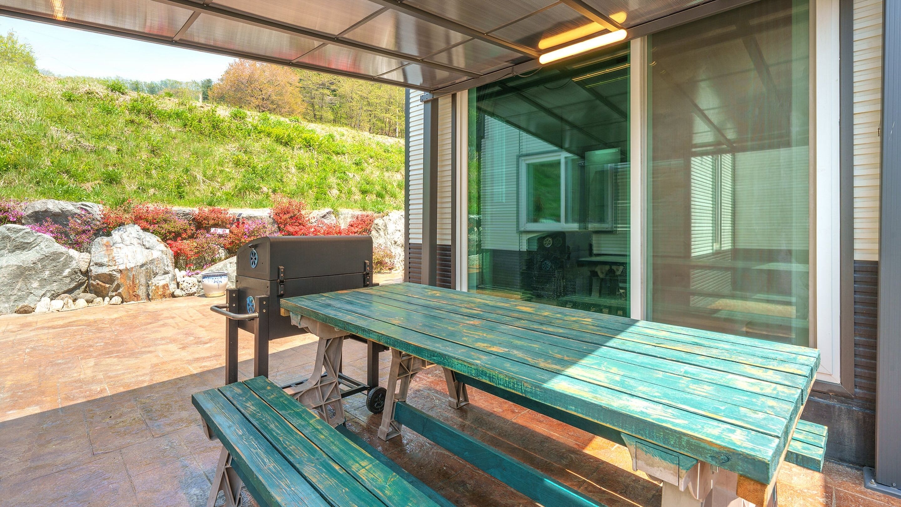 Terrassa exterior amb taula i bancs de fusta, una barbacoa i vista a un jardí paisatjista.