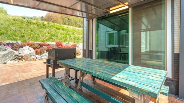 An outdoor patio with a wooden table and benches, a barbecue grill, and a view of a landscaped garden. - Hongcheon Pension 150 (Hongcheon)
