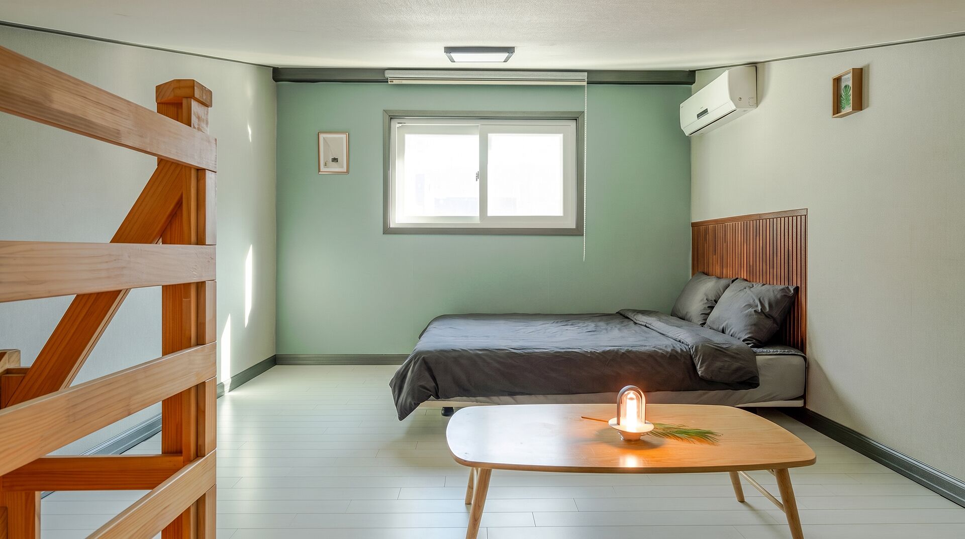 A bedroom with a bed, a bedside table with a candle, a window, and a wooden staircase.