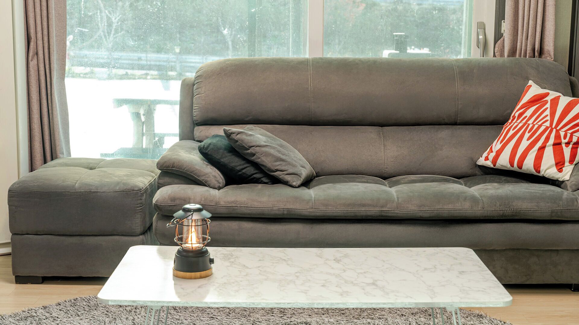 Un canapé gris agrémenté de plusieurs coussins et d’un oreiller rouge et blanc, une table basse en verre avec un pied en métal, et une petite lanterne posée sur la table.