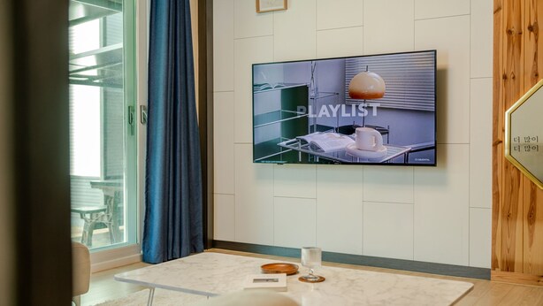 Une chambre d’hôtel moderne dotée d’un grand téléviseur à écran plat fixé au mur, qui affiche une image intitulée « LISTE DE LECTURE », d’une petite table avec un verre, un livre et un sous-verre en bois, ainsi que d’une fenêtre avec un rideau bleu.