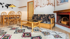 Un salon comprenant un canapé, une table basse en verre, une table à manger avec des chaises, une carte du monde accrochée au mur, une cheminée et un tapis coloré.