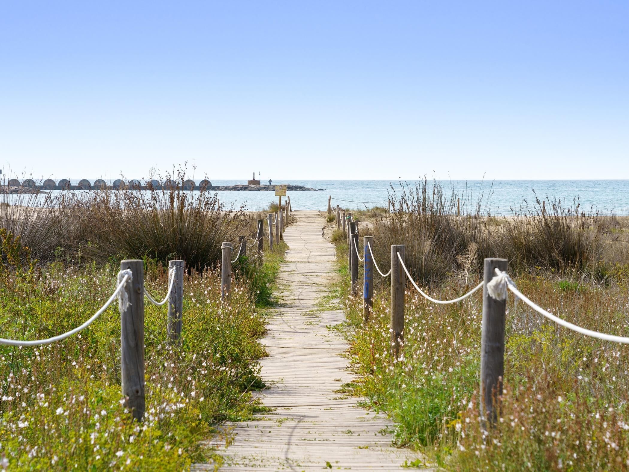 A wooden boardwalk leading to a beach with a clear view of the ocean and a distant pier.