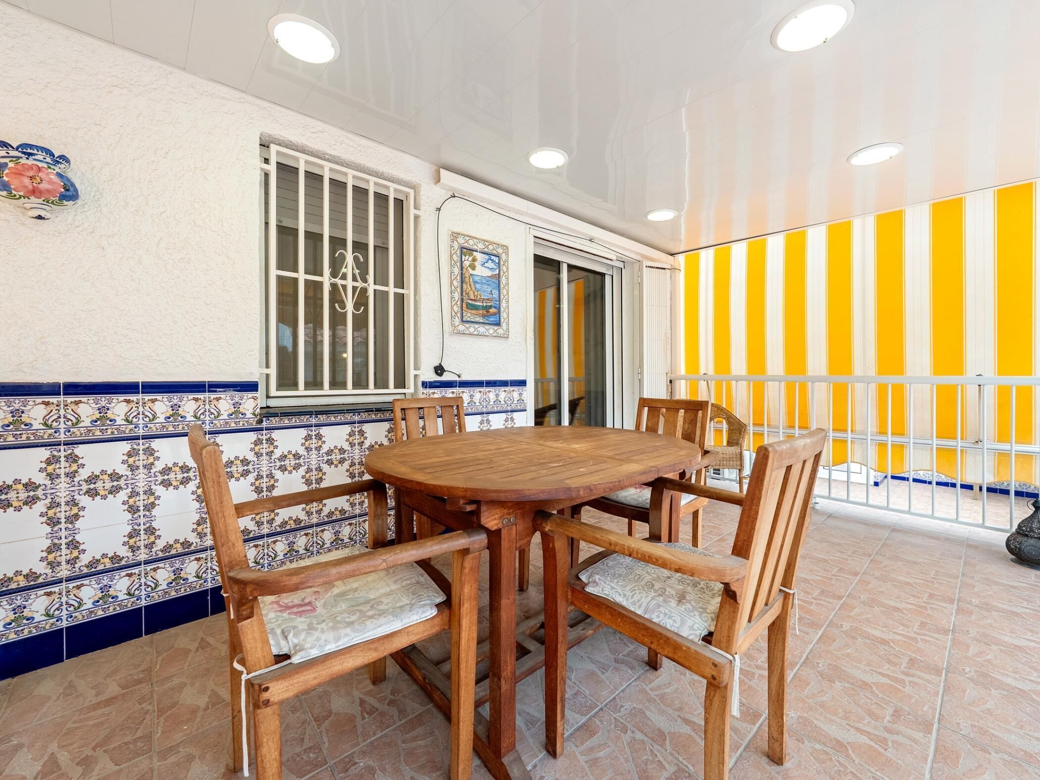 Een eetruimte met een houten tafel en stoelen, een betegelde muur met blauw-witte patronen, een raam met tralies en een gestreepte muur in geel en wit.