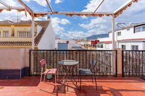 Een overdekt terras met een tafel en stoelen, een bamboescherm en uitzicht op omliggende gebouwen en bergen.