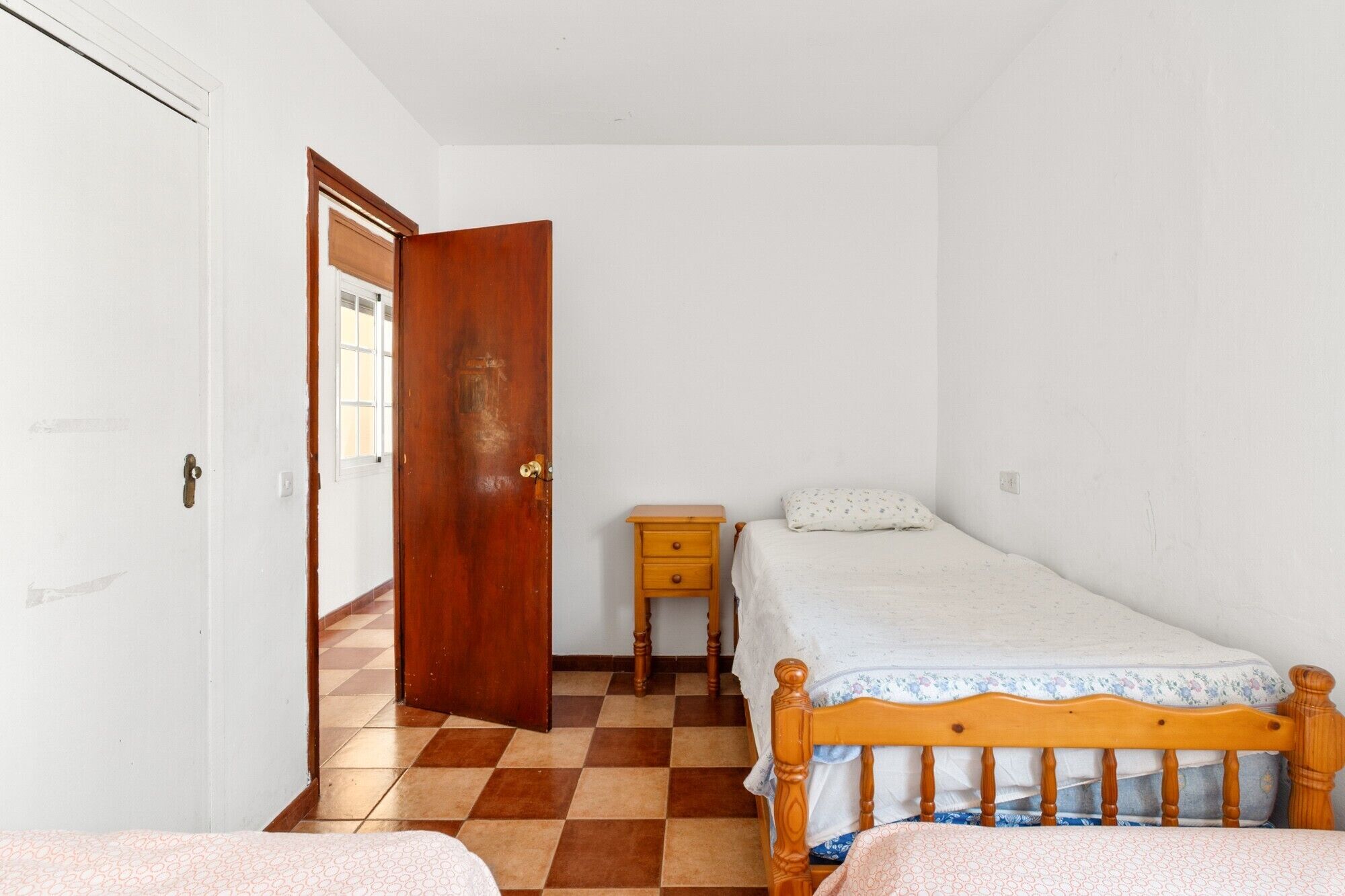 A bedroom with a checkered floor, a wooden bed, a nightstand, and a door.