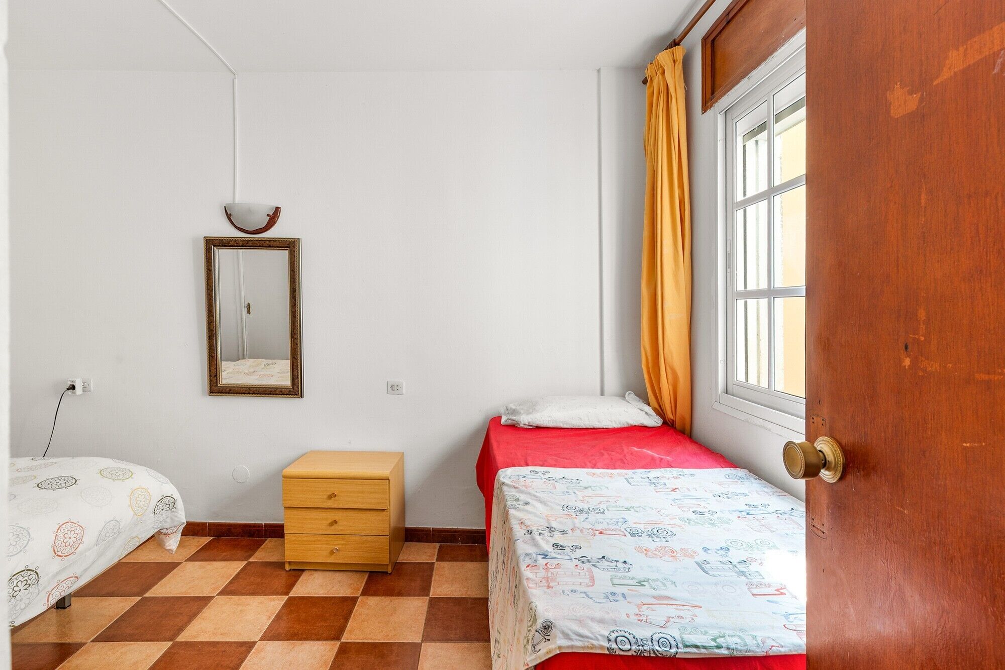 A bedroom with a checkered floor, a wooden nightstand, a bed with a patterned bedspread, a bedside table with a lamp, and a window with curtains.