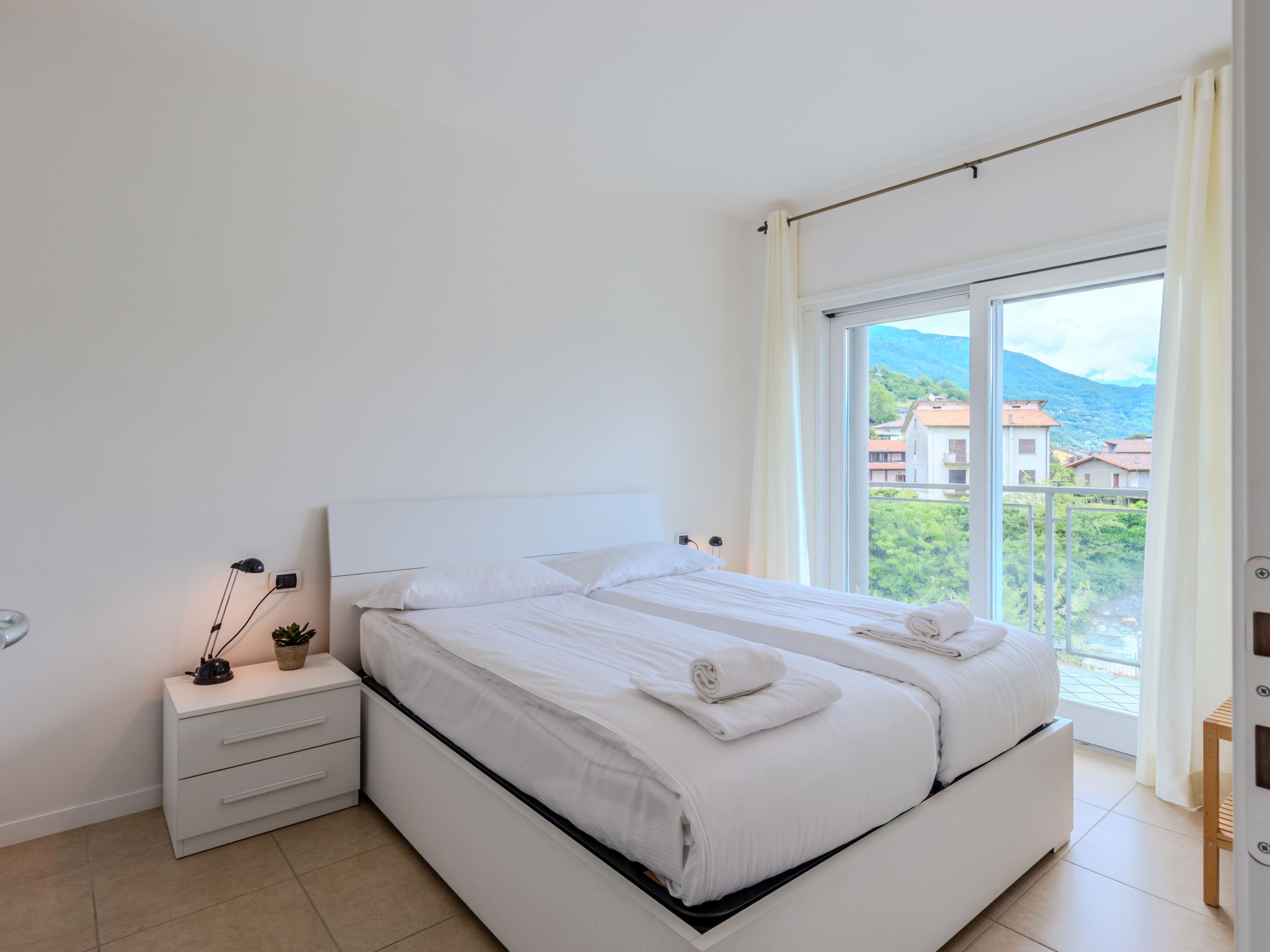 A modern bedroom with a large bed, a bedside table with a lamp, and a view of a mountainous landscape through a large window.