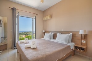 A hotel room with a large bed, a nightstand, a lamp, an air conditioning unit, and a view of the outdoors.