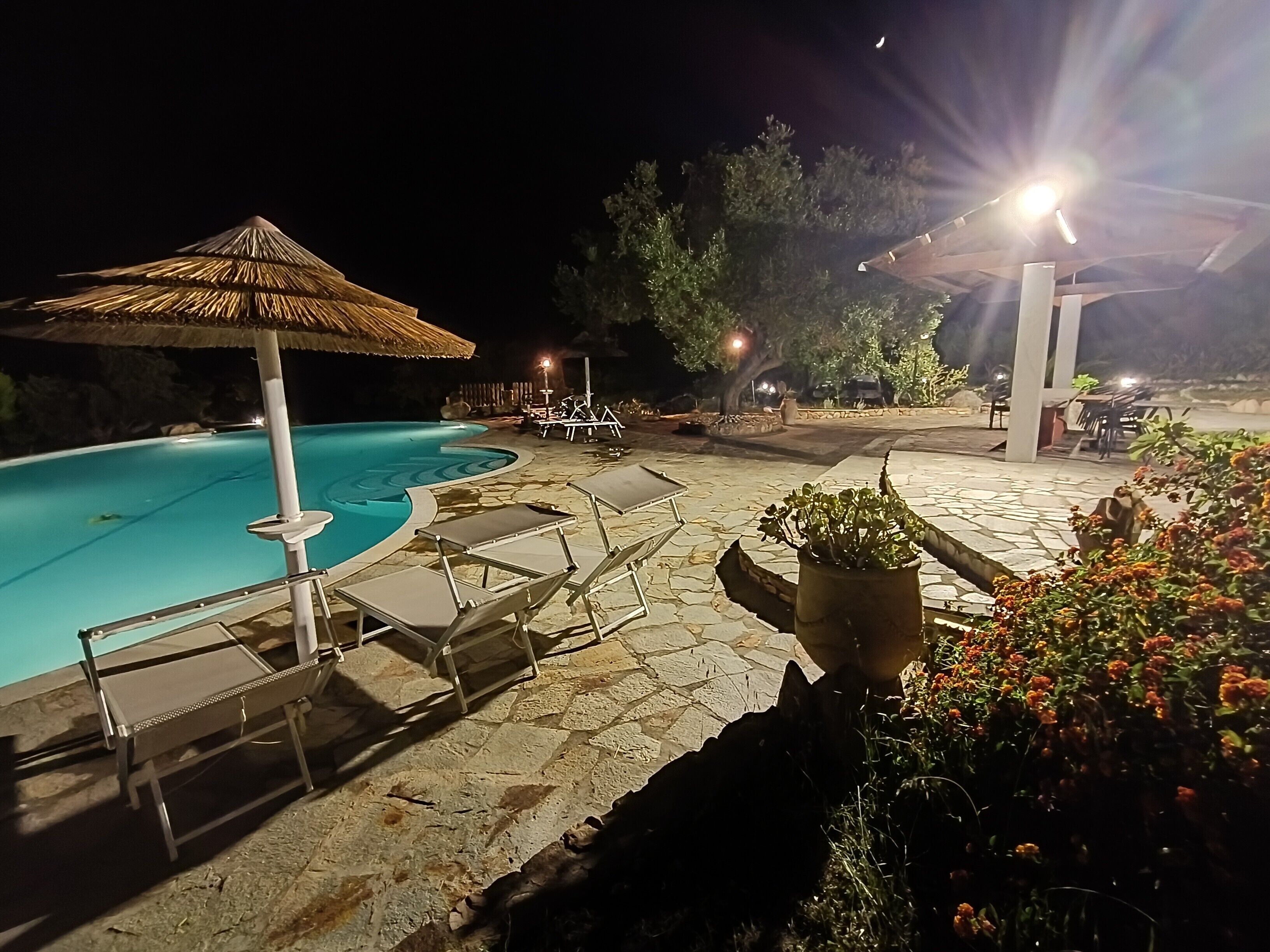 Un área de piscina nocturna con sillones de descanso, una sombrilla de paja y plantas en macetas.