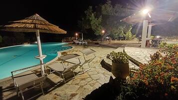 Un área de piscina nocturna con sillones de descanso, una sombrilla de paja y plantas en macetas.