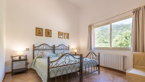 A bedroom with a metal bed frame, a bedside table with a lamp, a window with a view of greenery, and a radiator. - Room 'Campus' with Sea View, Wi-Fi and Air Conditioning (Villasimius)