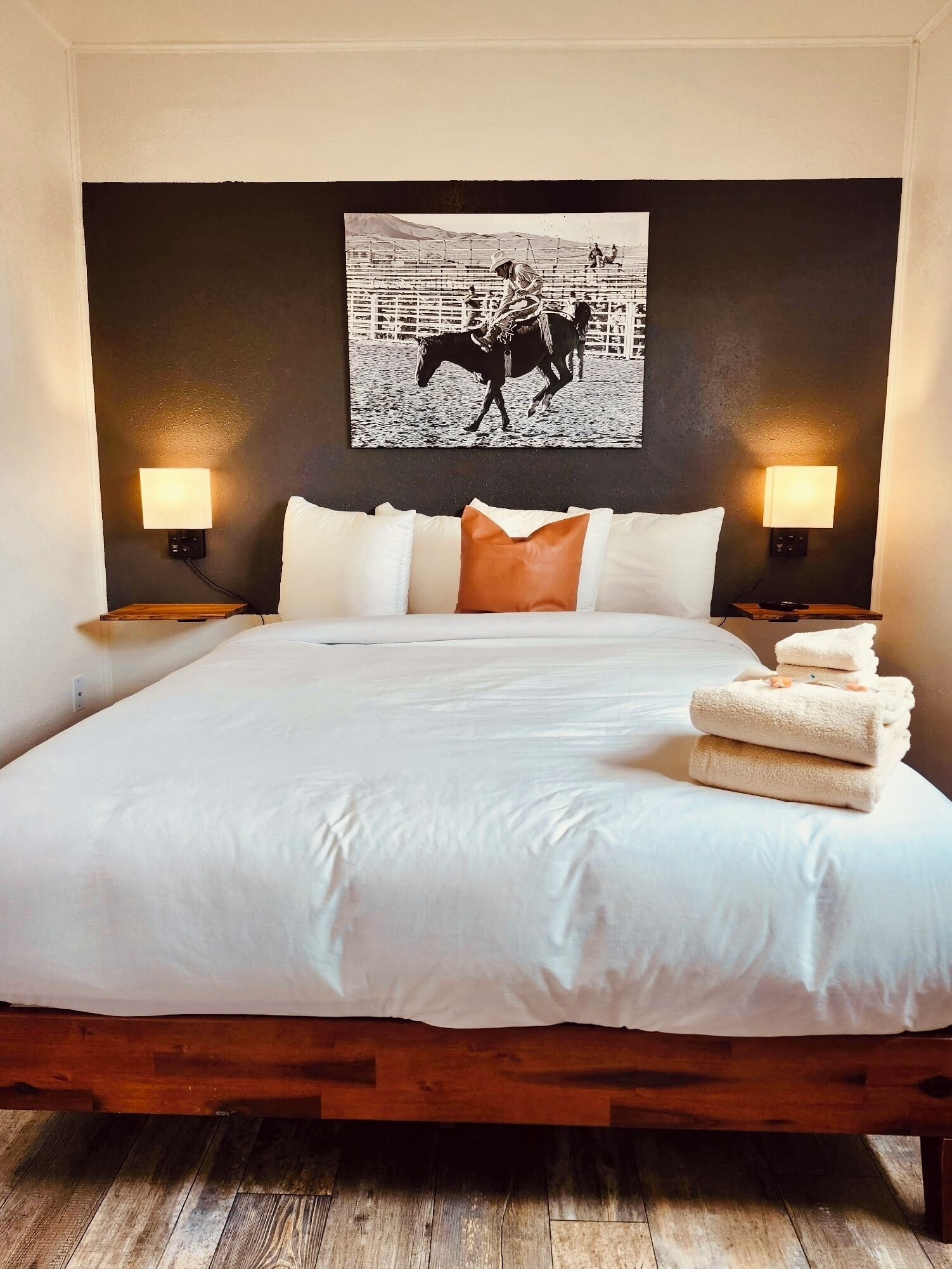 Un dormitorio con una cama grande, dos mesitas de noche con lámparas, un cuadro colgado de una carrera de caballos y suelo de madera.