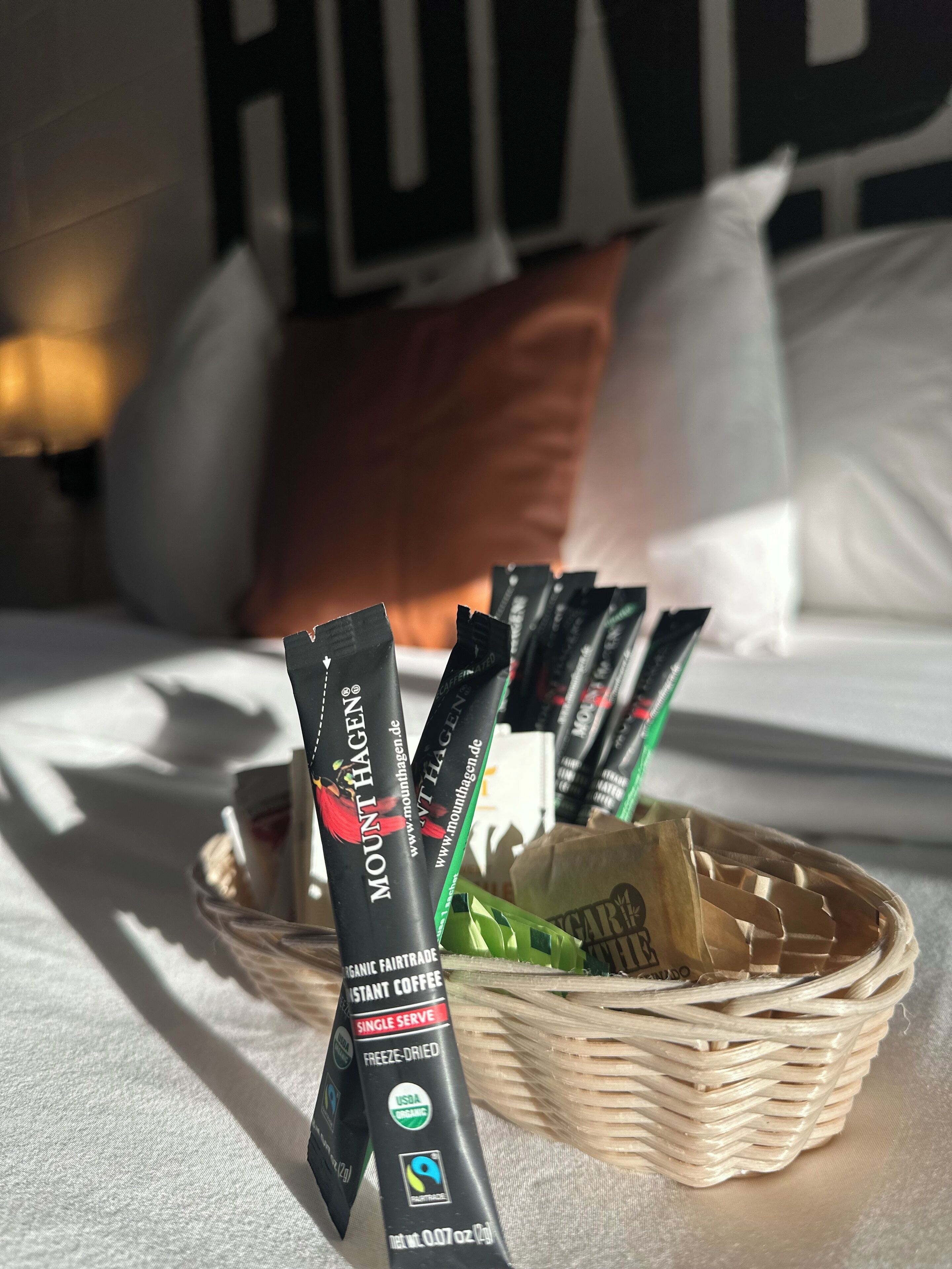 Una cesta con sobres de café instantáneo sobre una cama con ropa blanca y una almohada al fondo.