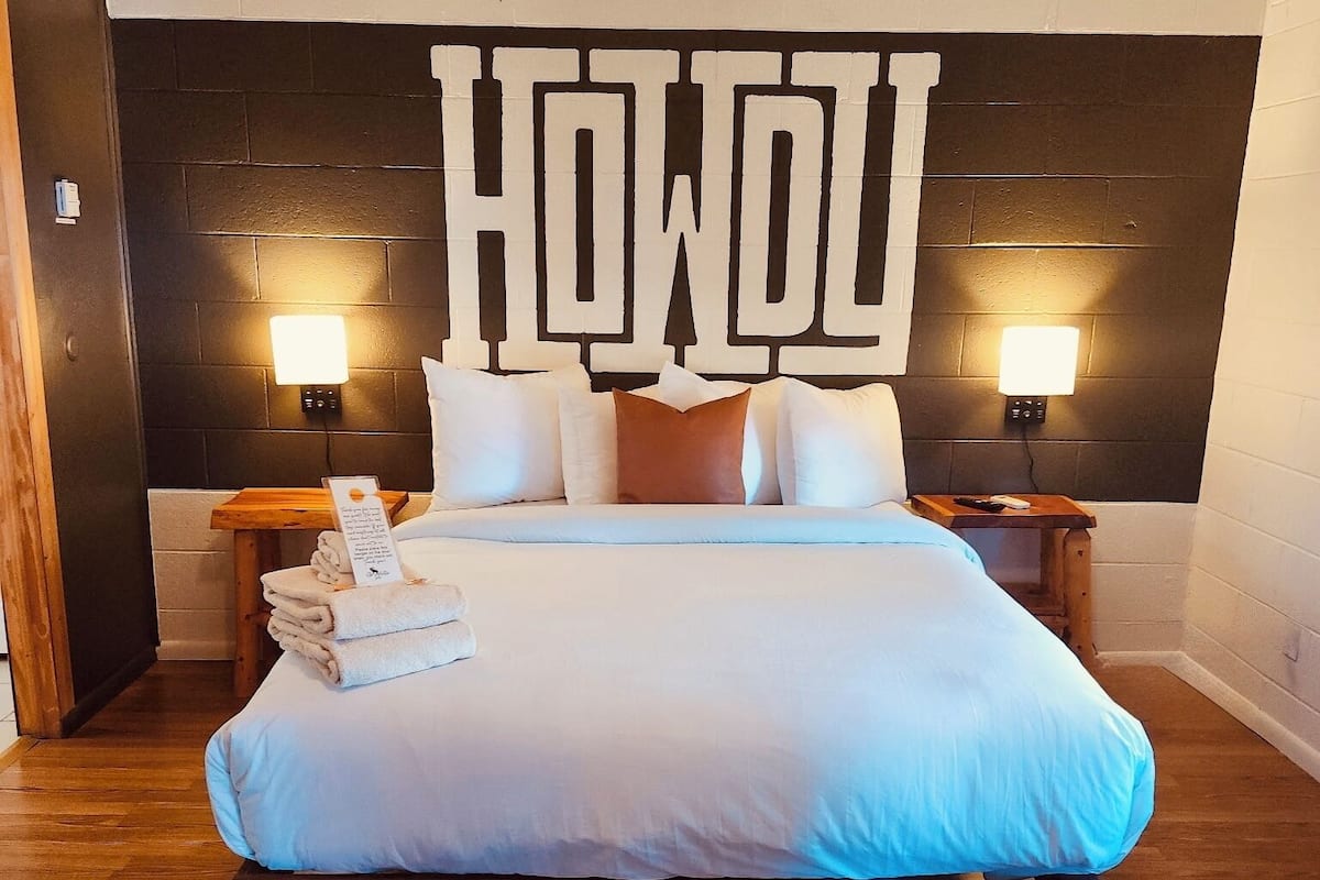 A hotel room with a bed, pillows, a nightstand, a wall-mounted light, and a towel stack.