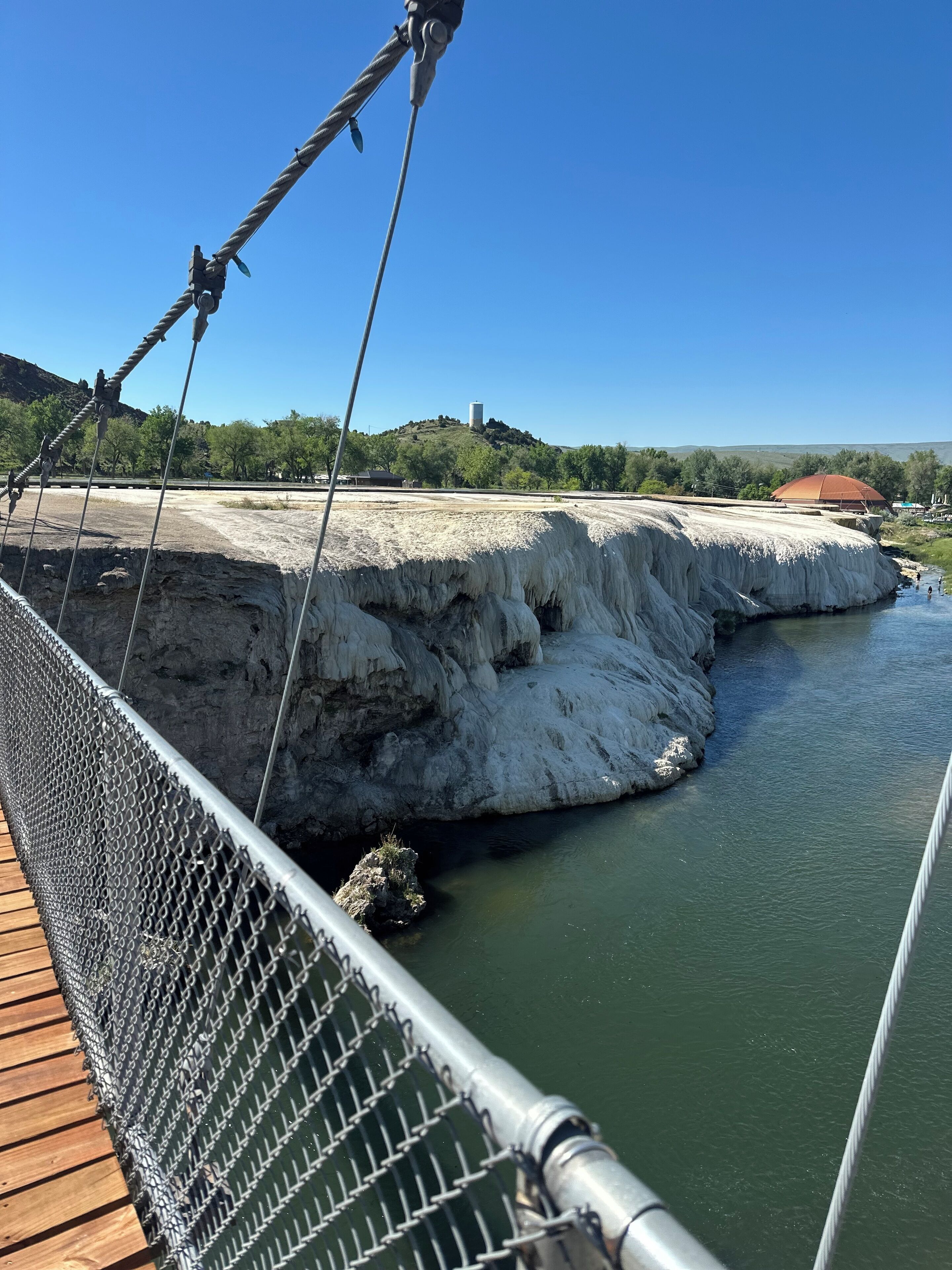A suspension bridge with a metal railing and a wooden deck, overlooking a river and a rocky landscape with a building in the background.