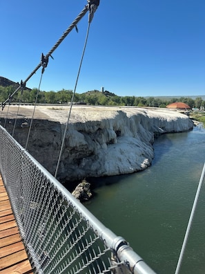 A suspension bridge with a metal railing and a wooden deck, overlooking a river and a rocky landscape with a building in the background. - Elk Antler Inn (Thermopolis)