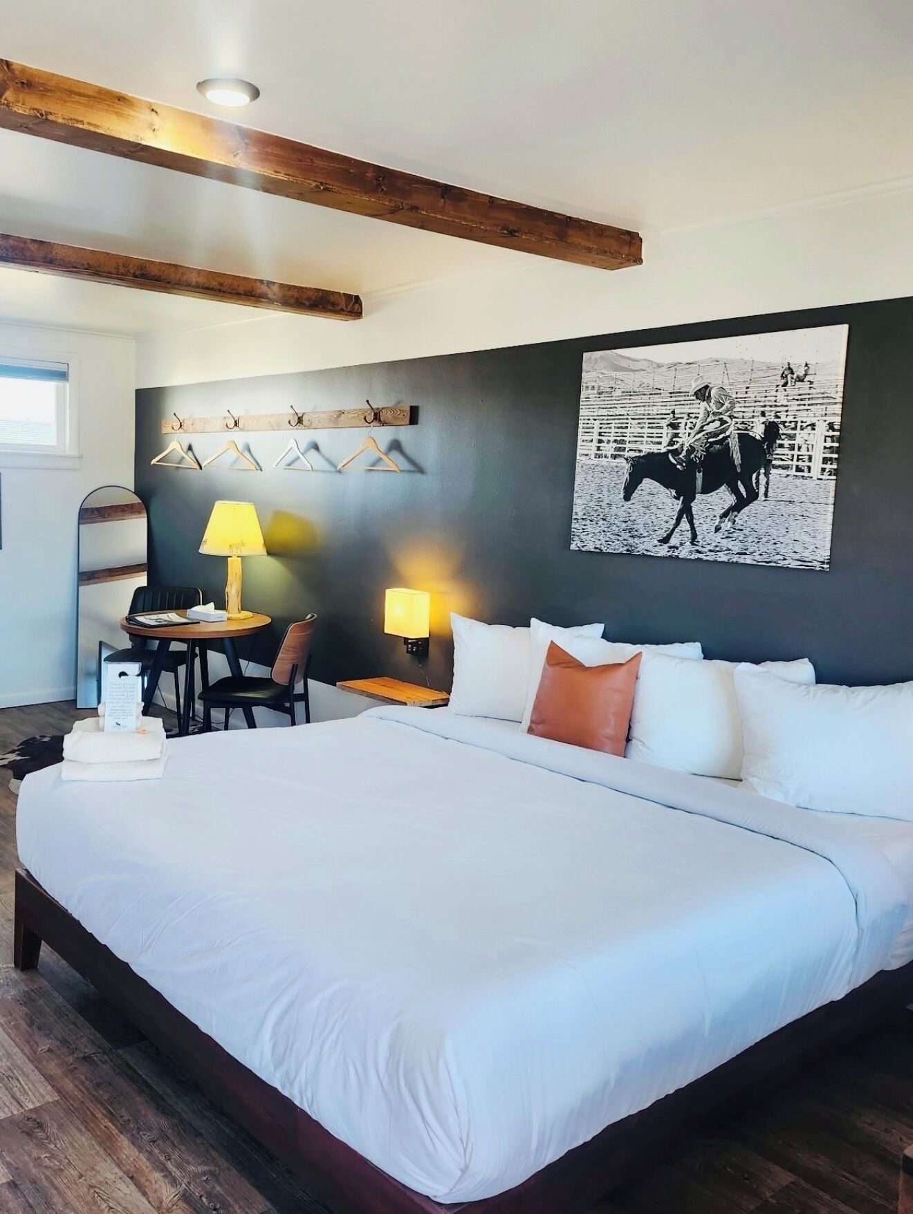 A hotel room with a large bed, a desk with a chair, a lamp, a coat rack, and a wall-mounted artwork.