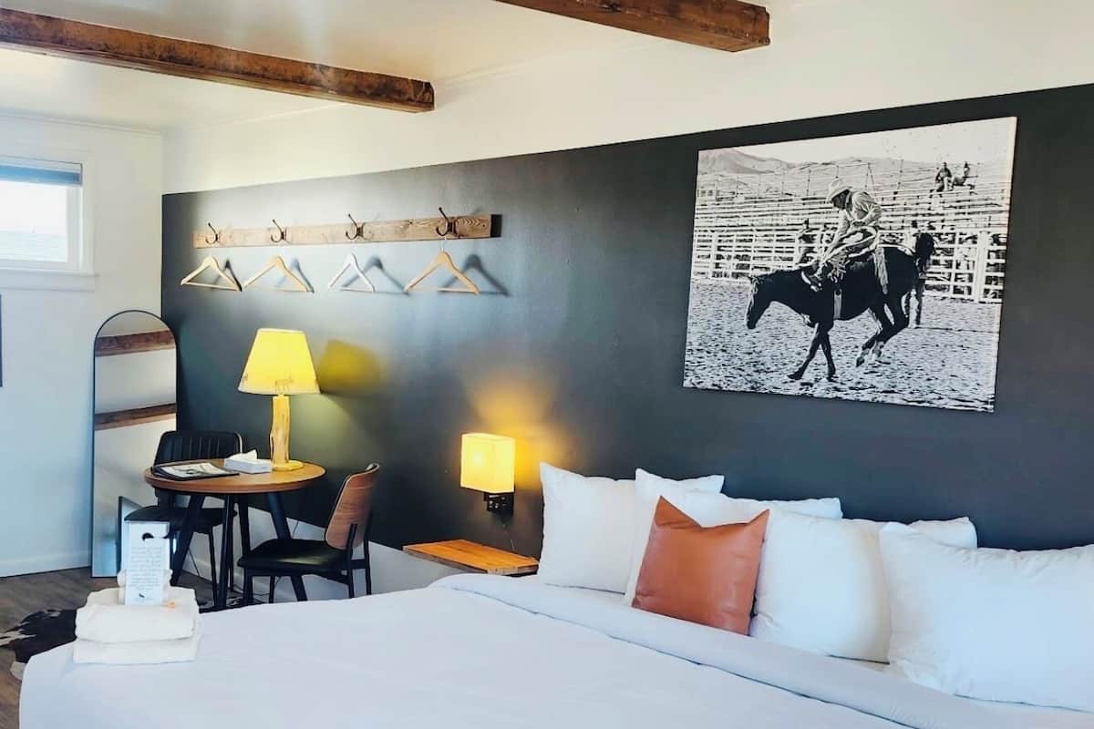 A hotel room with a large bed, a desk with a chair, a lamp, a coat rack, and a wall-mounted artwork.