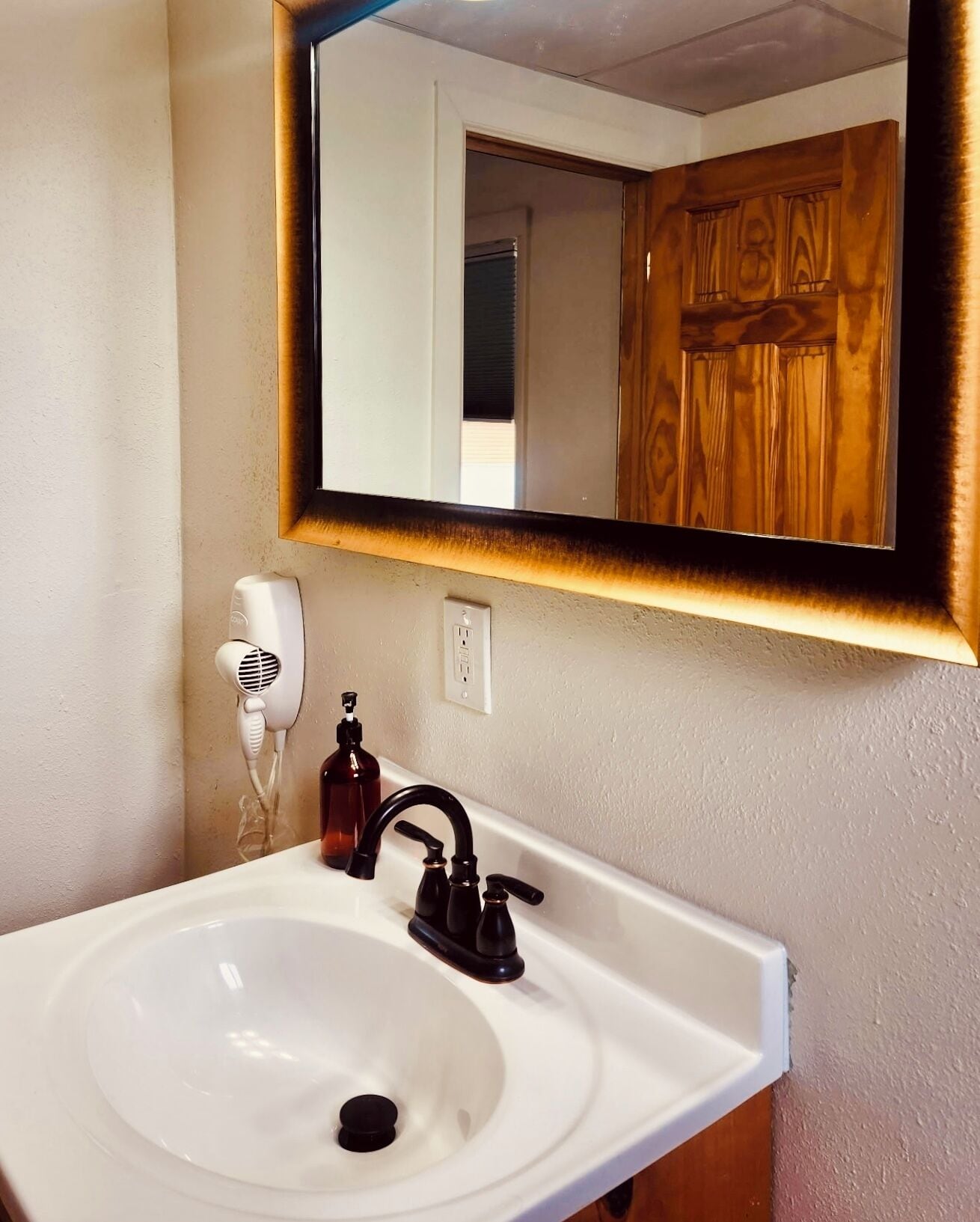 Un bagno con un lavandino bianco, uno specchio con cornice in legno, un asciugacapelli e un dispenser per sapone.
