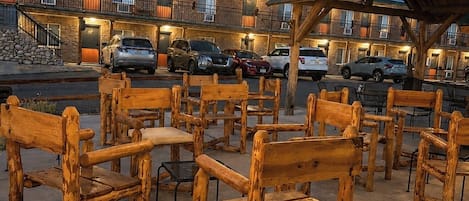 A rustic outdoor seating area with wooden benches and tables, a covered picnic area, and a building with multiple balconies and parked cars.