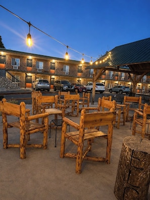 A rustic outdoor seating area with wooden benches and tables, a covered picnic area, and a building with multiple balconies and parked cars.