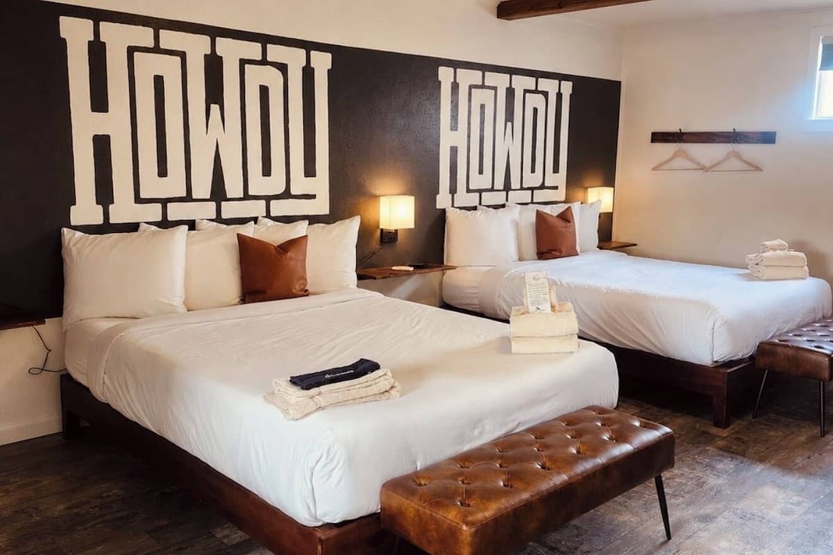 A hotel room with two beds, a large wall mural, wooden flooring, and a bench.