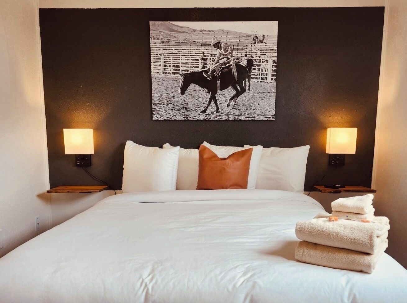 A hotel room with a large bed, two bedside lamps, a mounted picture of a horse rider, and a stack of towels on the bed.