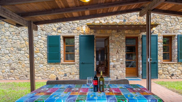 A stone building with a covered patio, a table with a colorful tablecloth, and two bottles of wine.