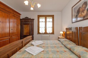 Una camera da letto con un letto, un armadio in legno, una cassettiera, una finestra con tende, un termosifone e un quadro appeso al muro.