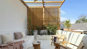 Un área de estar al aire libre con un diseño moderno, que incluye una pérgola de madera, asientos acolchados y mesas de madera redondas.