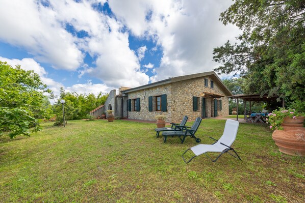Une maison en pierre avec une pelouse, deux chaises longues et une pergola.