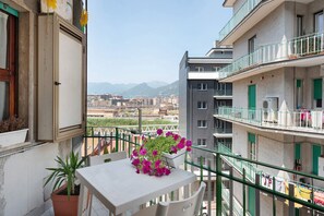 Un balcón con mesa, plantas en macetas y vistas a edificios y montañas.