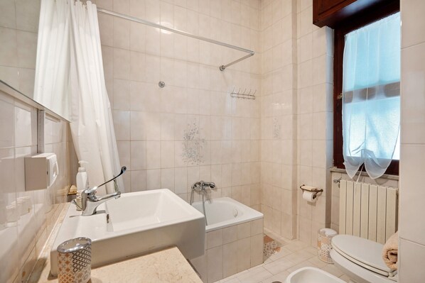 Un baño con bañera, ducha, inodoro y ventana con cortina.