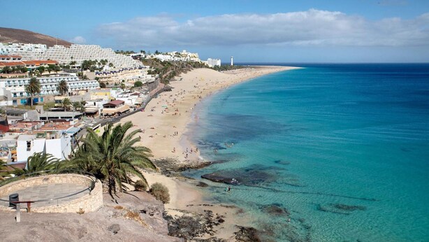 A coastal town with a sandy beach, clear blue waters, and buildings along the shore.