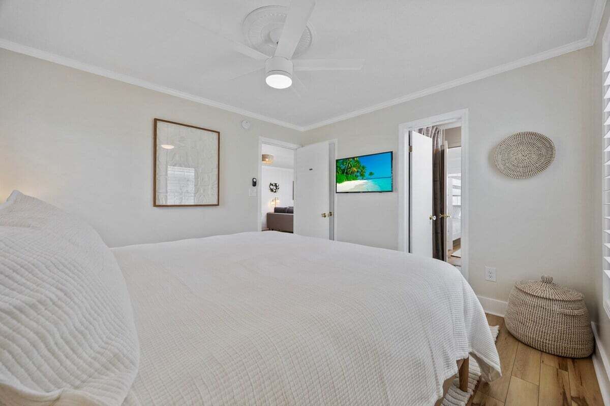A bedroom with a large bed, a ceiling fan, a framed picture, a television, and a woven basket.